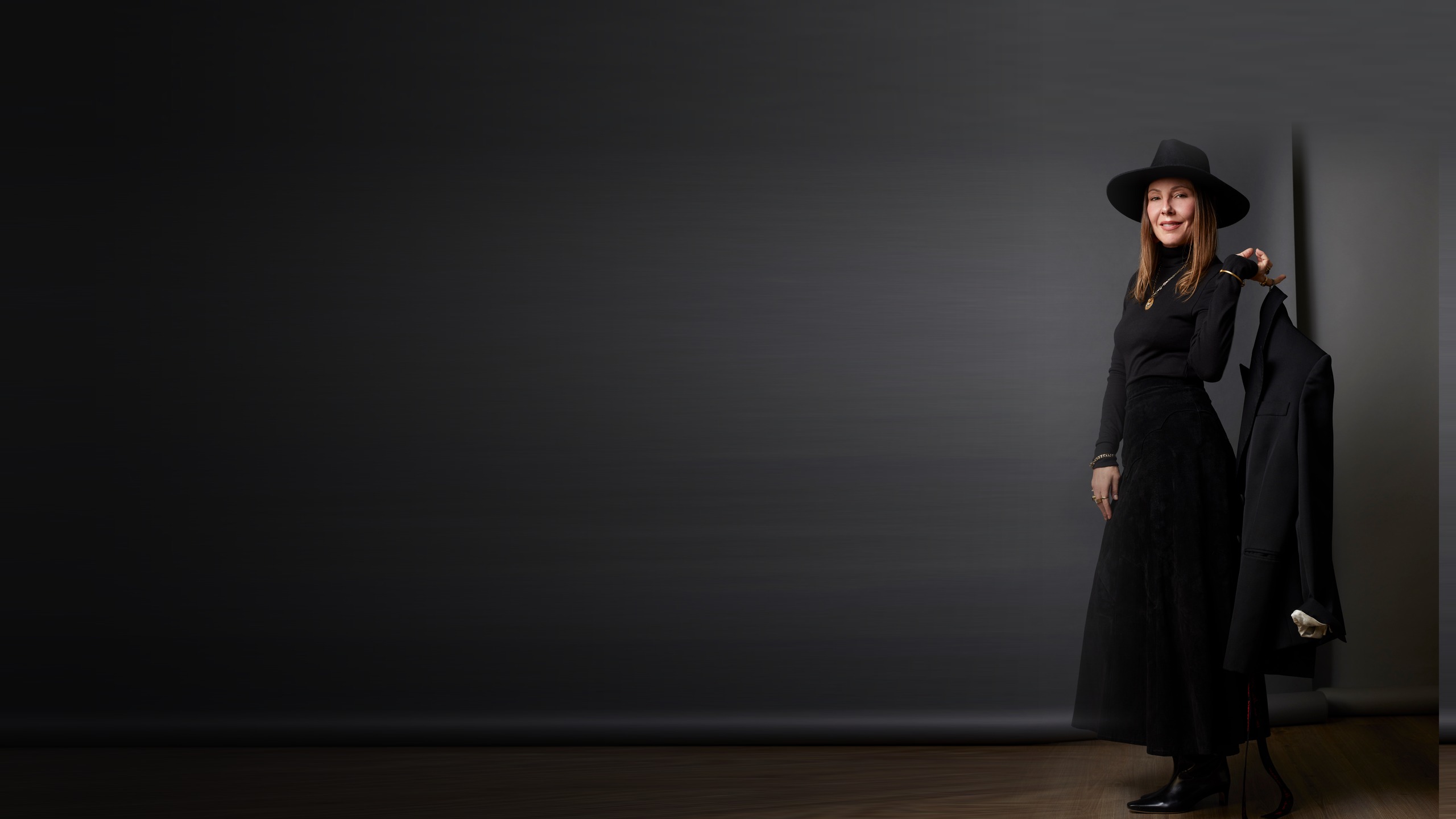 Gara Gambucci in black, holding a tailored jacket, against a dark studio wall.